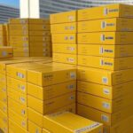 Caixas de vacinas contra febre amarela em centro de distribuição no Distrito Federal, com arquitetura de Brasília ao fundo.