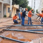 Rua vazia na Asa Norte de Brasília com tubulações e placa da Caesb indicando corte de água, ao fundo edifícios governamentais.