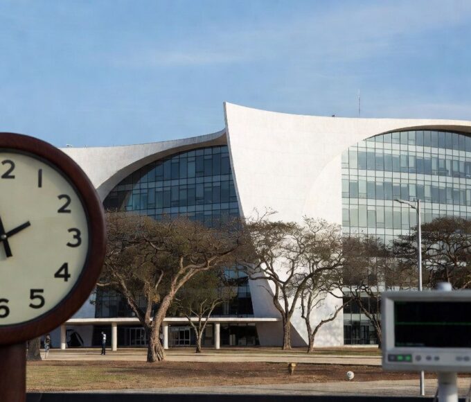Edifício da Câmara Legislativa do DF em Brasília com relógio parado e monitor cardíaco, simbolizando lentidão em lei para ecocardiograma em bebês com Down e riscos cardíacos.