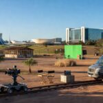 Polo de cinema no DF com edifícios e equipamentos de filmagem em área de assentamento rural, arquitetura de Brasília ao fundo.